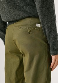 Olivové nohavice s textúrovanou tkaninou, s zadnou vreckom so zapínaním na gombík a so značkovou etiketou vyšitou nad vreckom.