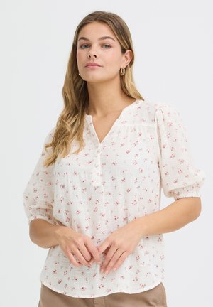 Kvinde med langt blondt hår, der bærer guldhoop-øreringe og en hvid bluse med rød floral print og puffede ærmer.