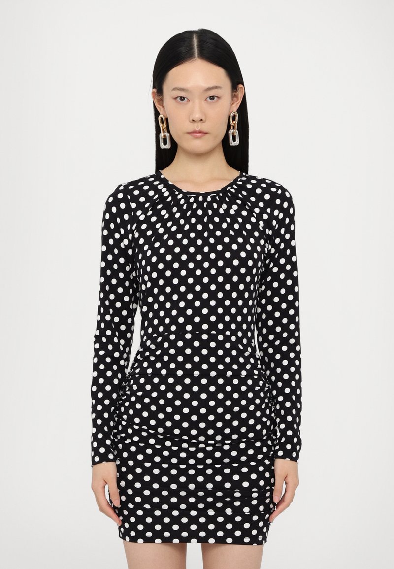 Black dress with white polka dots, long sleeves, gathered neckline, and fitted silhouette, featuring a ruched skirt detail.
