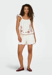 Top in lino bianco con spalline arricciate e ricamo floreale rosso, abbinato a pantaloni corti bianchi con orlo scallopato accentuato da cuciture rosse.