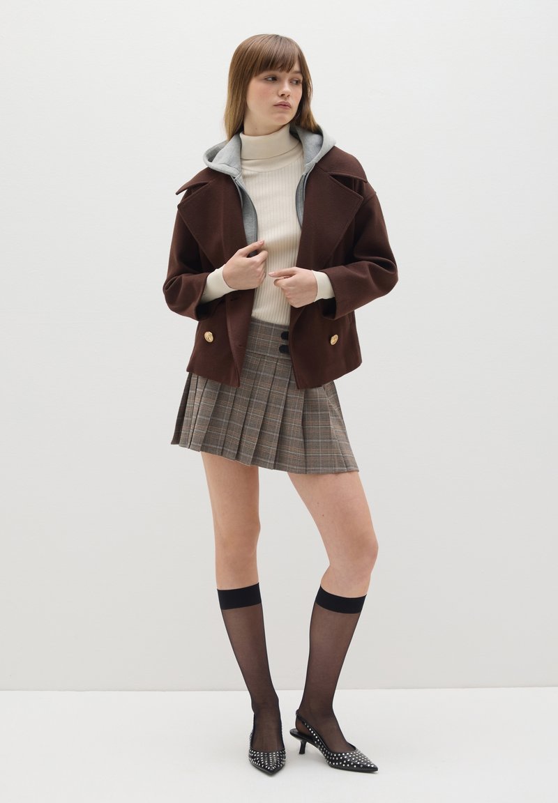Chaqueta marrón de doble botonadura con capucha gris, cuello alto de punto beige, falda corta de cuadros, medias altas negras y tacones con tachuelas.