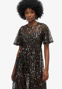 Vestido negro con mangas cortas y transparentes, adornado con filas verticales de lentejuelas metálicas. Tejido texturizado y fluido con escote redondo.