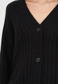 Cardigan noir en maille avec motif structuré, encolure en V et deux boutons noirs visibles sur la partie supérieure du torse d'une personne.