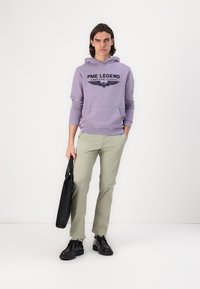 Svetlofialová mikina s čiernym textom a grafikou krídla, v kombinácii so svetlozelenými nohavicami a čiernymi topánkami, drží čiernu tašku.