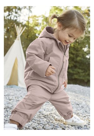 Chaqueta con capucha de color rosa claro con cierre de cremallera, combinada con pantalones a juego. Textura suave y puños elásticos, sobre un fondo de piedras.