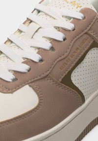 Gros plan d'une basket beige et blanche avec des lacets blancs, un panneau en cuir perforé, et "REPLAY DENIM LOVERS" embossé sur le côté.
