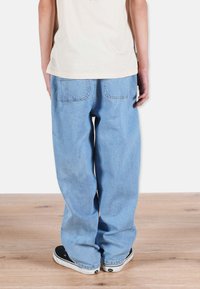 Vaaleansinisiä, väljiä farkkuja, joissa on kaksi takataskua, yhdistettynä vaaleanvihreään paitaan ja mustiin lenkkareihin, puulattialla.