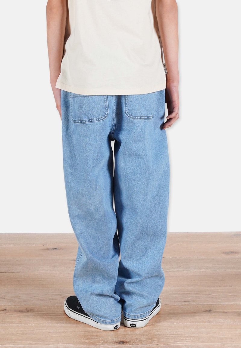 Vaaleansinisiä, väljiä farkkuja, joissa on kaksi takataskua, yhdistettynä vaaleanvihreään paitaan ja mustiin lenkkareihin, puulattialla.