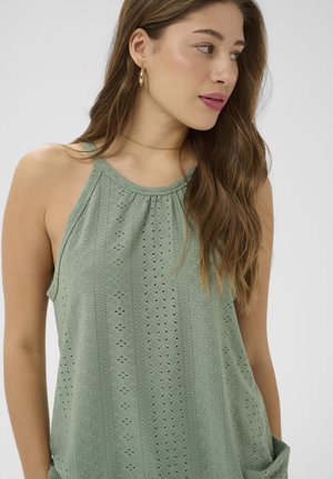 Mladá žena s dlhými hnedými vlasmi, ktorá nosí zlaté kruhové náušnice a svetlozelený vyšívaný tielkový top, pozerá sa doprava.