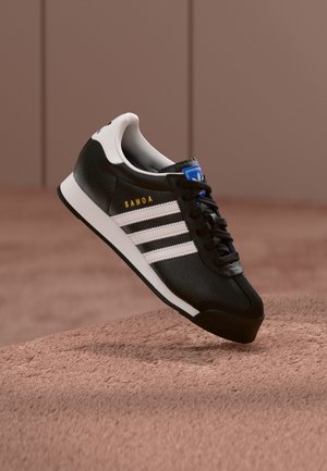 Baskets Adidas Samoa noires avec des bandes blanches et un col de talon, lacets noirs et étiquette bleue sur la languette flottant au-dessus d'une surface texturée.