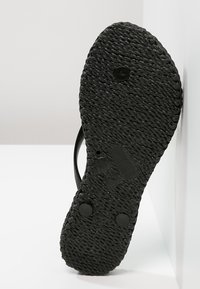 Musta flip-flop-pohja, jossa on teksturoitu, kudottu kuvio. Kaksi pyöreää kumipäällystettä tarjoavat pito. Kantapää on hieman kohotettu, minimalistinen muotoilu ilman koristeita.