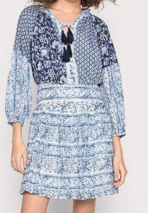 Žena v modrobílých květovaných šatech s třásněmi na výstřihu a dlouhými nadýchanými rukávy stojí před jednobarevným pozadím.