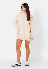 Vestido sudadera beige con un bolsillo delantero y puños acanelados. El modelo lo combina con zapatillas blancas con rayas negras. Diseño sencillo y casual.