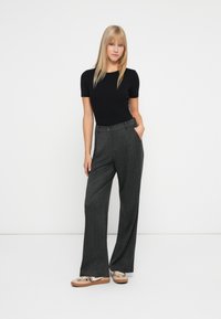 Vrouw met steil blond haar, gekleed in een zwart shirt met korte mouwen, grijze wijde broek en beige sneakers met panterprint accenten.