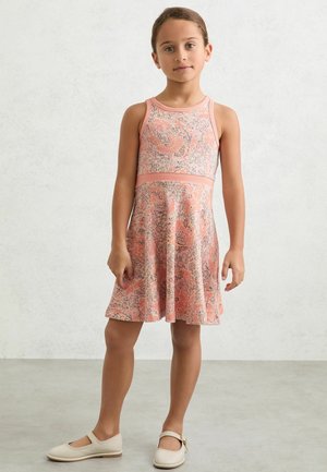 Fiatal lány áll, ujjatlan rózsaszín virágos ruhát és bézs Mary Jane cipőt visel, egyszerű világos háttér előtt.