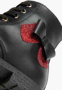 Schwarzer Lederschuh mit runder Zehenpartie, schwarzen Schnürsenkeln und glitzerndem roten Schleifenakzent. Texturierte Sohle ist am unteren Rand sichtbar.