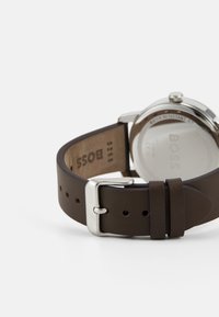 Reloj con correa de piel marrón y caja de acero inoxidable plateado. Cuenta con una textura suave, hebilla de metal y nombre de la marca grabado en la parte posterior.