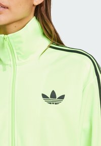 Neon-grüne Zip-up-Jacke mit schwarzen Streifen auf den Schultern und Logo. Glatte Textur, hoher Kragen und taillierte Passform.