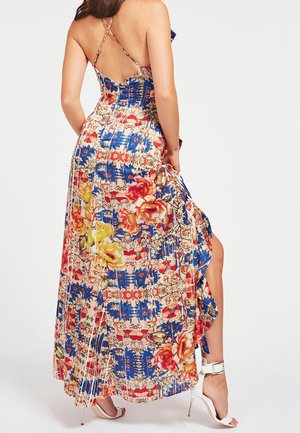 Kobieta w kolorowej, kwiatowej sukience maxi na cienkich ramiączkach z rozcięciem po boku, ubrana w białe sandały na wysokim obcasie.