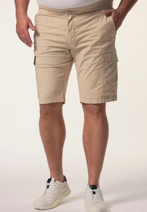Mand iført beige cargoshorts med sidelommer og hvide sneakers, stående mod en ensfarvet lys baggrund.