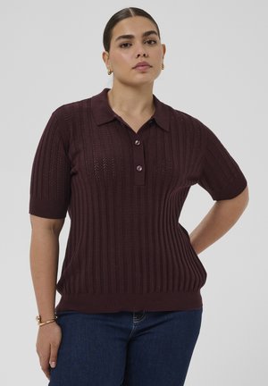 Vrouw met donker haar in een knot, gekleed in een bordeauxrode polo van gebreide stof met korte mouwen en een blauwe spijkerbroek, staand met één hand in haar zij.