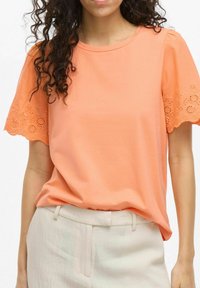 Top de manga corta en algodón color coral con detalles de encaje en las mangas, escote redondo y corte holgado, combinado con pantalones de colores claros.