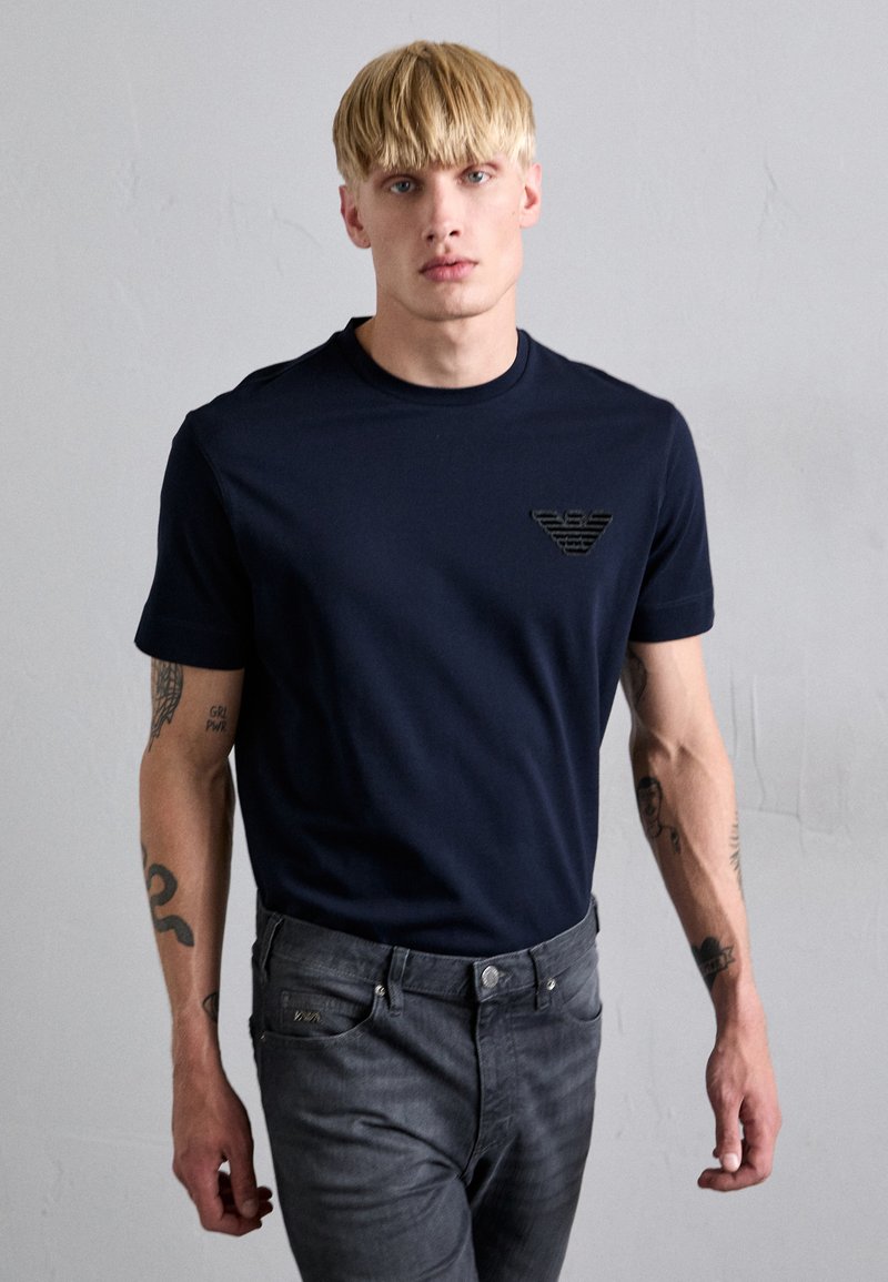 Marineblaue Baumwoll-T-Shirt mit kleinem schwarzem Logo auf der linken Brust, kombiniert mit dunkelgrauen Jeans. Model mit kurzem blonden Haar.