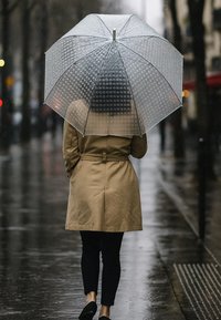 Clear, dome-shaped umbrella with a textured surface. Brown trench coat, dark pants, and black shoes; wet pavement reflects surrounding lights.