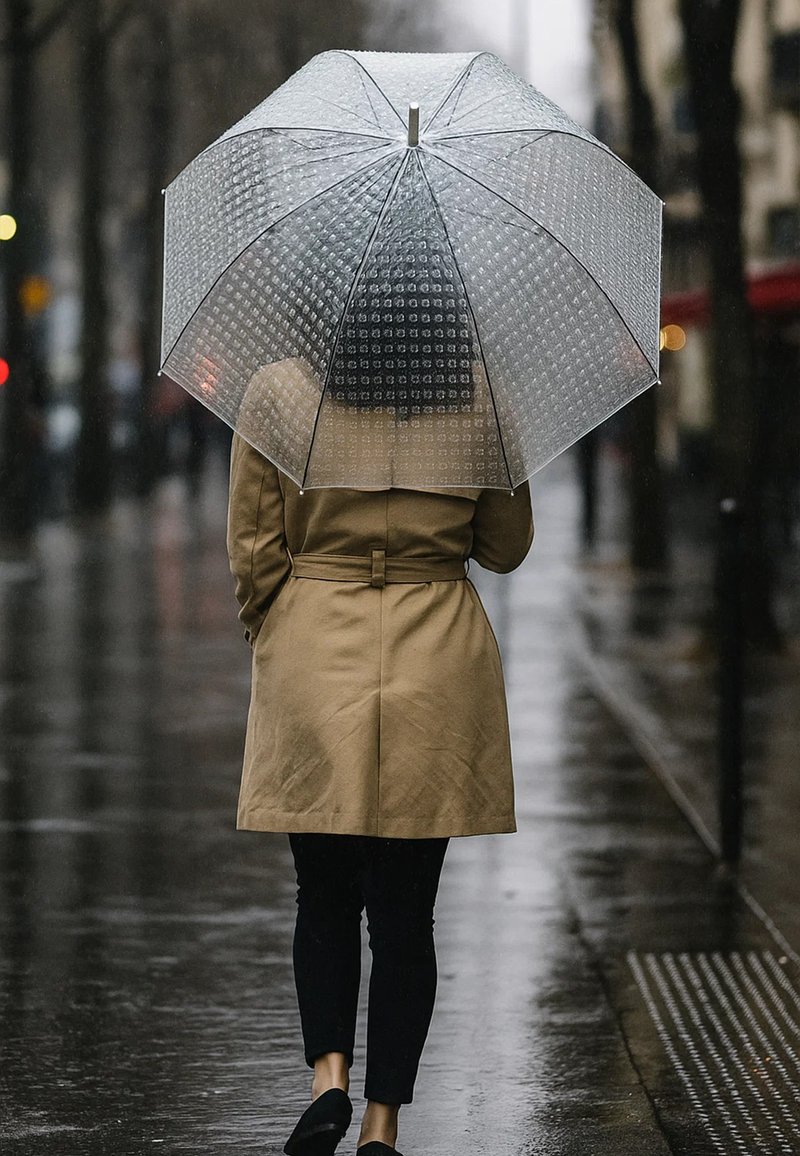 Clear, dome-shaped umbrella with a textured surface. Brown trench coat, dark pants, and black shoes; wet pavement reflects surrounding lights.