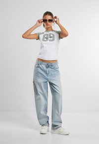 Witte crop top met groene mouwen en het nummer "89", gecombineerd met lichtblauwe baggy jeans en witte sneakers. Het model draagt een zonnebril.