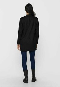 Cappotto lungo nero con taglio dritto, caratterizzato da un colletto alto, abbinato a jeans scuri e stivali neri chunky. Texture del tessuto liscia.