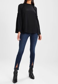 Schwarze Bluse mit langen Ärmeln und gerafften Details an den Schultern, kombiniert mit dunkelblauen Jeans, die seitliche Schlitze am Saum aufweisen, und schwarzen Stiefeln mit Absatz.