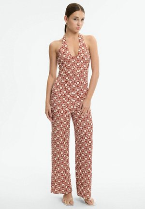 Mujer de pie con las manos relajadas, vestida con un mono sin mangas estilo halter con un estampado de conchas en rojo y blanco y sandalias con correas transparentes.