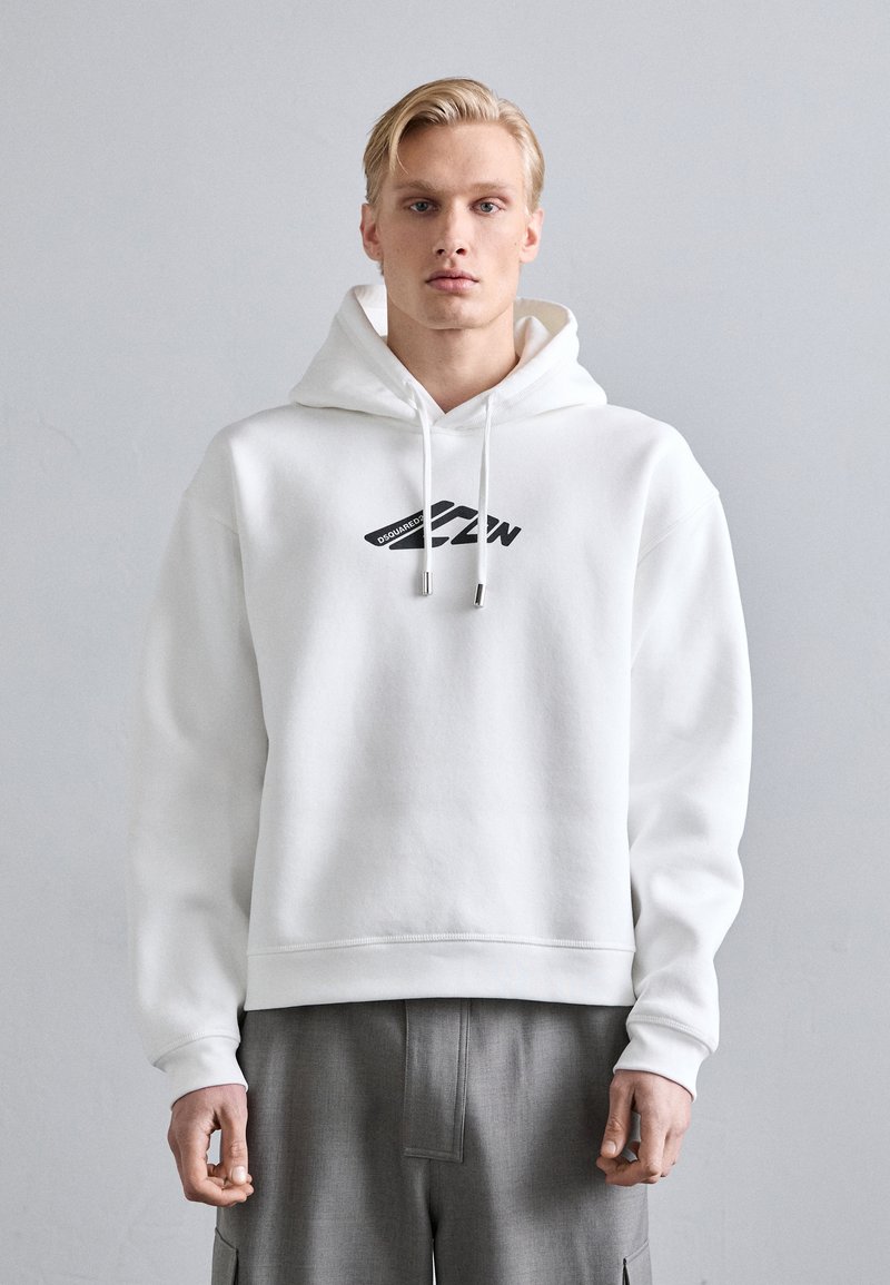 Sudadera blanca de algodón con un logotipo gráfico en la parte frontal, que cuenta con una capucha con cordón y puños acanalados. Llevada sobre pantalones grises con pliegues.