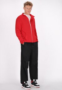 Rode ritssluitingjas met hoge kraag, gecombineerd met losse zwarte broek en zwart-witte sneakers met rode accenten. Iedereen heeft een gladde textuur.