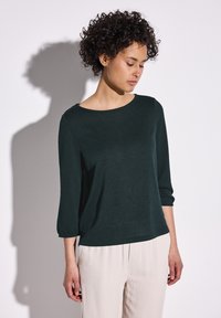 Haut à manches longues vert foncé, avec un col rond et une coupe décontractée, fabriqué à partir d'un tissu texturé. Associé à un pantalon de couleur claire.