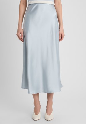 Femme portant une jupe midi en satin bleu clair et des talons blancs à bout pointu, debout devant un fond clair uni.