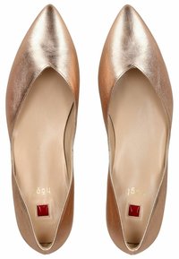 Płaskie buty w kolorze różowego złota z ostrym noskiem, z wycięciem o zaokrąglonym kształcie i małym czerwonym prostokątnym akcentem przy pięcie. Gładka faktura.