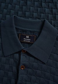 Temno modra pletena polo majica z reliefnim karo vzorcem, rjavimi gumbi in etiketo Matinique Heritage Apparel znotraj ovratnika.