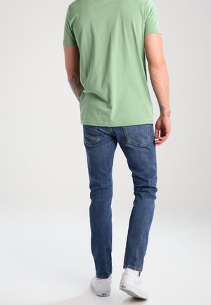 Man draagt een groen kortärmelig shirt, blauwe spijkerbroek en witte sneakers, staat met de rug naar de camera tegen een effen lichte achtergrond.