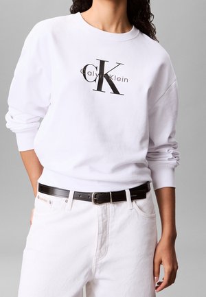 Mujer vistiendo sudadera blanca de Calvin Klein con gran logo negro "CK", jeans blancos de talle alto y cinturón de cuero negro, posando contra un fondo gris.