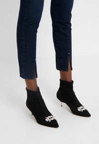 Botines de ante negros con puntera afilada, que presentan adornos decorativos de strass y un tacón delgado. Combinados con jeans oscuros con dobladillos abiertos.
