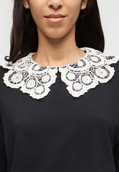 Femme portant un haut noir avec un large col en dentelle blanche présentant des motifs circulaires et floraux.