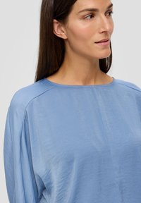 Helles blaues, locker sitzendes Oberteil mit runder Halslinie, langen Ärmeln und einer glatten, leicht glänzenden Oberfläche. Minimale Nähte und Details.