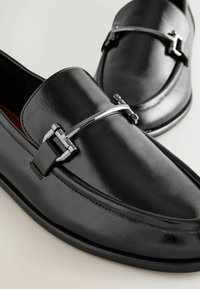 Mocassins en cuir noir avec détail boucle en métal argenté, finition polie, bouts arrondis et talons bas sur fond clair.