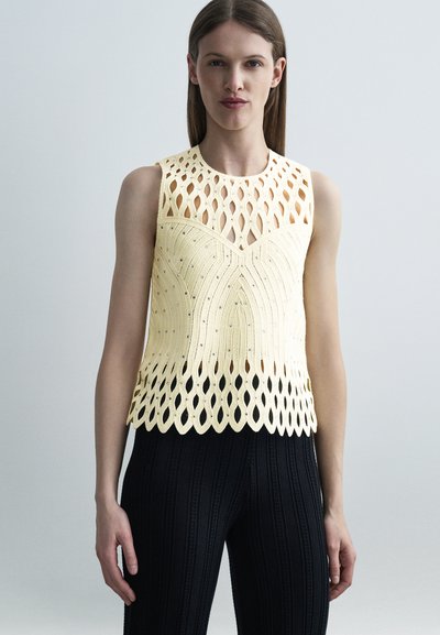 Femme portant un haut en maille jaune pâle sans manches avec un motif ajouré en boucle et un pantalon texturé noir sur fond uni.