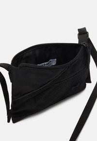 Černá látková crossbody taška s hlavním zipovým oddílem a síťovaným akcentem. Obsahuje plochý popruh pro nošení. Vnitřní etiketa je viditelná.