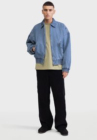 Denimjack met een relaxte pasvorm, lichtblauwe kleur, rits aan de voorkant, elastische boorden en zijzakken, gedragen over een beige T-shirt en donkere broek.