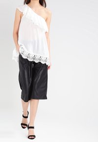 Top blanco de un solo hombro con borde de volantes y detalle de encaje, combinado con culottes de piel sintética negra y tacones negros de tiras.