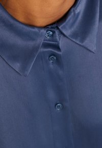 Satijnen blauwe kraagshirt met een knoopsluiting, voorzien van afgeronde kraagranden en een soepele textuur, detail van twee zichtbare knopen.